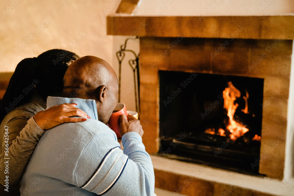 Senior African couple by cozy fireplace drinking hot chocolate together - Elderly people hugging each other inside cabin home - Soft focus on woman hand Senior African couple by cozy fireplace drinking hot chocolate together - Elderly people hugging each other inside cabin home - Soft focus on woman hand