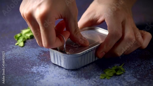 Hand opening a can of mackerel on a dark blue table with parsley. Ready to eat.