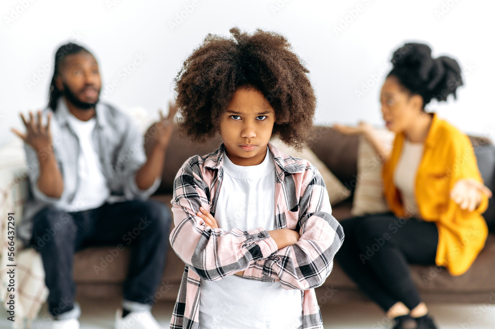 Obraz premium Difficulties in the family. An African American unhappy preschool girl looks offendedly at the camera, against the background, parents sort things out, shout at each other, swear, the child is scared