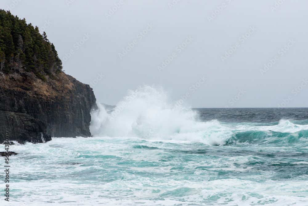 Large teal green colored waves crashed against jagged rocks along the shoreline. The rough angry sea forms waves and white spray and froth as it rolls in on the beach. The ocean is flowing quickly. 