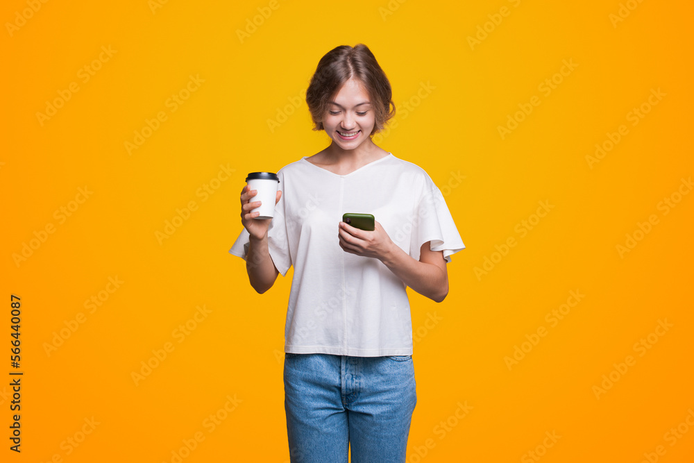 Attractive young brunette woman typing message, drinking takeaway coffee, using online mobile application isolated over yellow background Attractive young brunette woman typing message, drinking takeaway coffee, using online mobile application isolated over yellow background