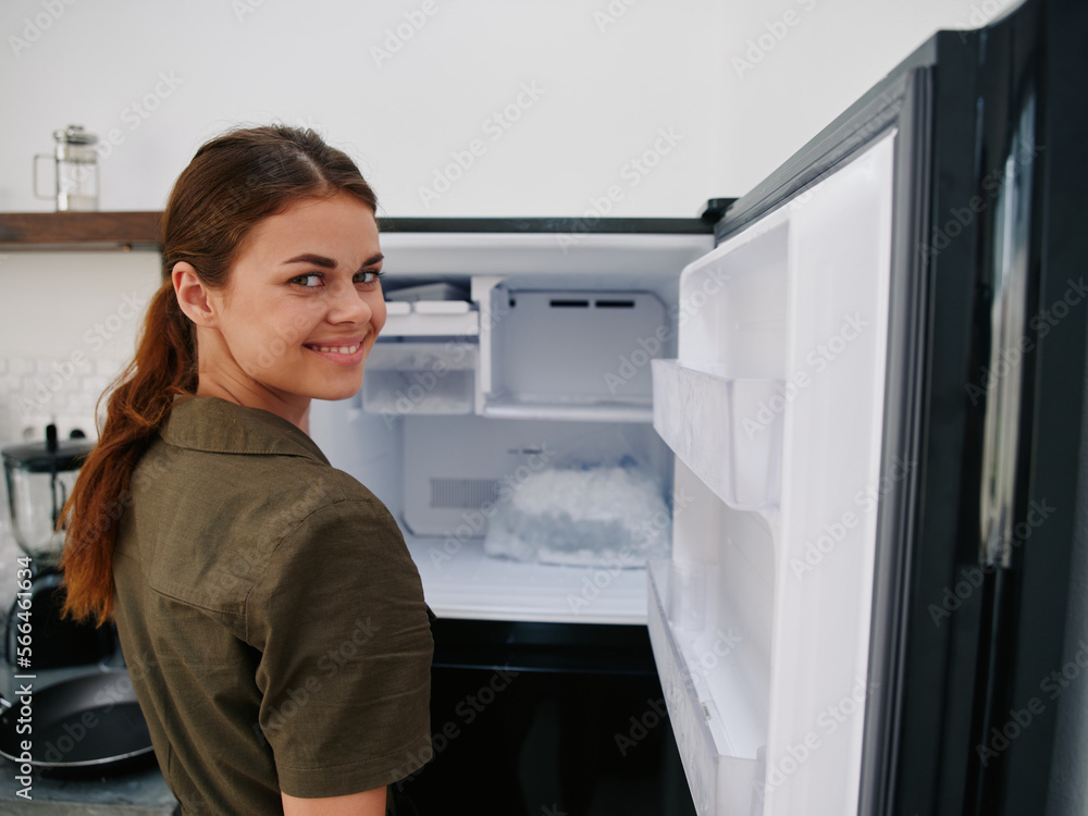 Woman smiling with teeth looking into camera in kitchen at home opened freezer empty with ice inside, home refrigerator, defrosted, view from back, stylish interior. Woman smiling with teeth looking into camera in kitchen at home opened freezer empty with ice inside, home refrigerator, defrosted, view from back, stylish interior.