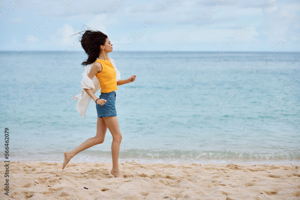 Sports woman runs along the beach in summer clothes on the sand in a yellow tank top and denim shorts white shirt flying hair ocean view, beach vacation and travel