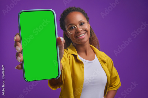 Young smiling African American woman student shows phone with green screen advertising application for self-education and gaining new knowledge through smartphone stands in purple studio. Soft focus