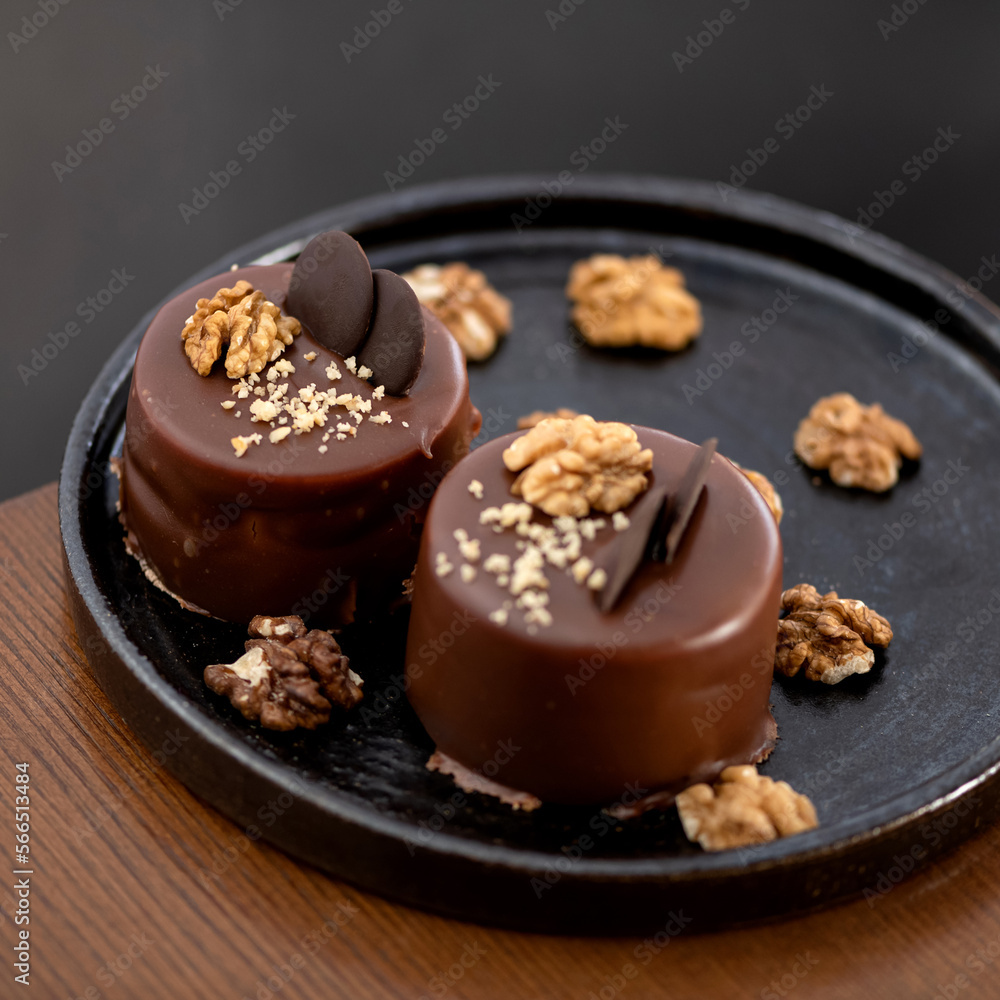 Two chocolate small desserts on plate. Cakes are decorated with nuts and chocolate glaze. Festive baking. Wooden table. View from above. Dark background. Close-up. Soft focus. Copy space. Two chocolate small desserts on plate. Cakes are decorated with nuts and chocolate glaze. Festive baking. Wooden table. View from above. Dark background. Close-up. Soft focus. Copy space.