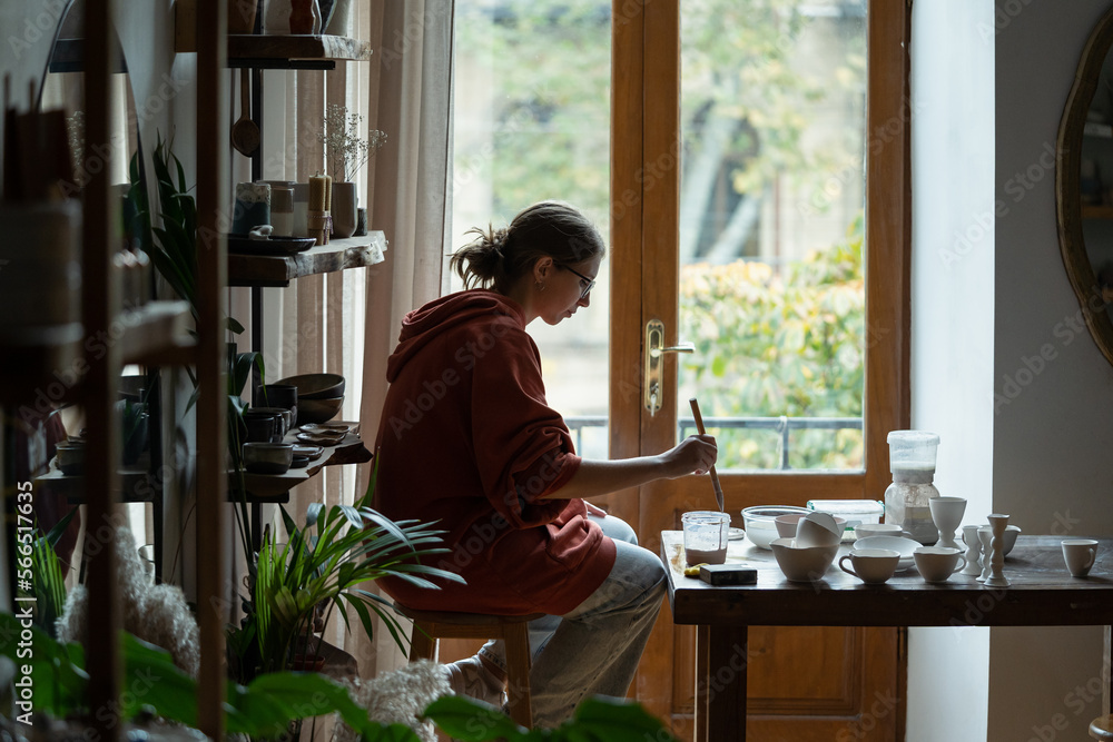 Thoughtful creative woman artist decorating clay plates after kiln firing sits at table in workshop. Introversion girl in casual clothes enjoys process drawing patterns on porcelain and ceramic dishes Thoughtful creative woman artist decorating clay plates after kiln firing sits at table in workshop. Introversion girl in casual clothes enjoys process drawing patterns on porcelain and ceramic dishes