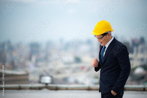 Young business man and architect or contractor in formal clothing with yellow hardhat standing with open arms wearing eyeglasses on rooftop of new building against tall buildings