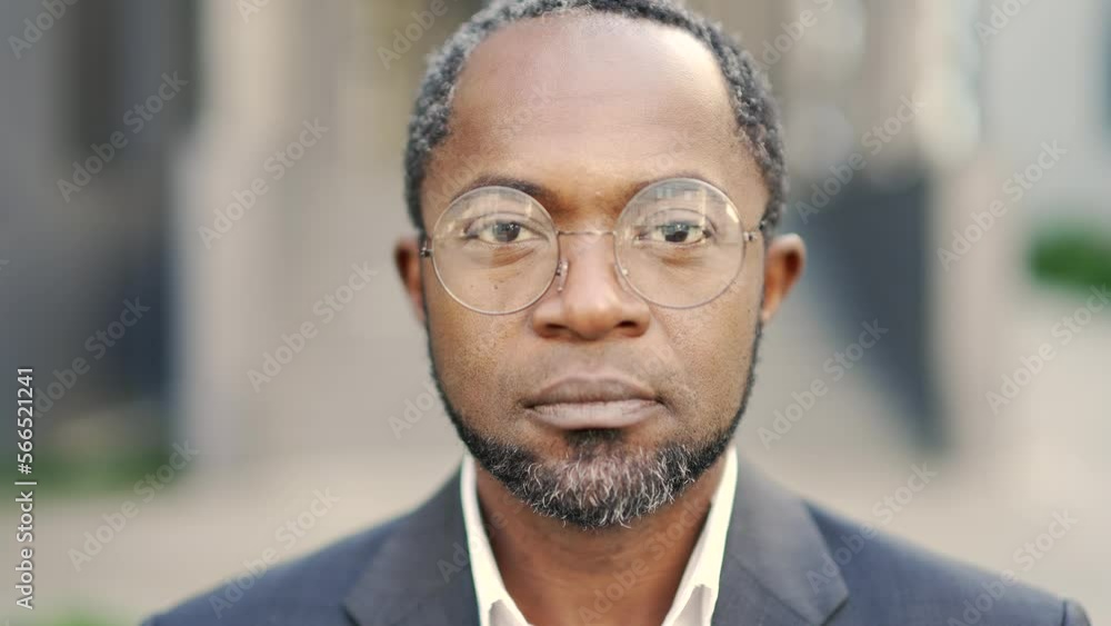 Close-up portrait of serious mature african american man wearing glasses looking at camera outdoors. Сonfident middle-aged businessman owner is standing in a formal suit in front of an office building Close-up portrait of serious mature african american man wearing glasses looking at camera outdoors. Сonfident middle-aged businessman owner is standing in a formal suit in front of an office building