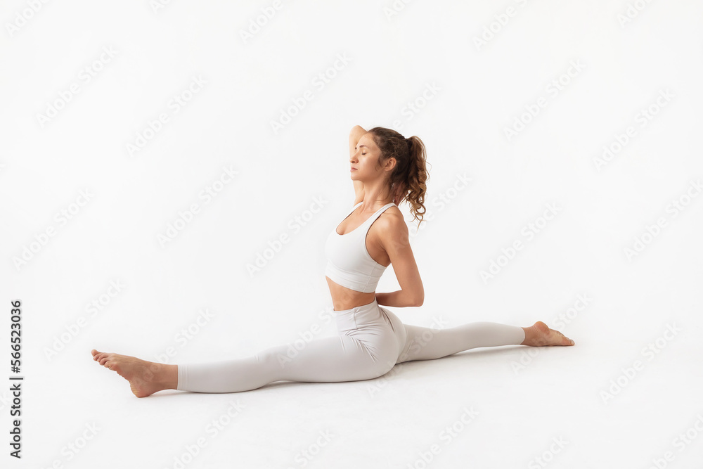 Young woman practicing yoga, doing a variation of the Hanumanasana exercise with hands behind her back, a pose with a longitudinal split, exercising in white sportswear on a light background Young woman practicing yoga, doing a variation of the Hanumanasana exercise with hands behind her back, a pose with a longitudinal split, exercising in white sportswear on a light background
