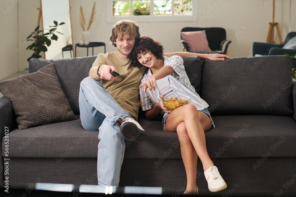 Young woman with bowl of potato chips sittingf on sofa next to her husband and pointing at tv screen while guy choosing channel Young woman with bowl of potato chips sittingf on sofa next to her husband and pointing at tv screen while guy choosing channel