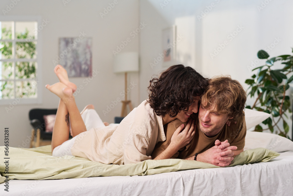 Happy young barefoot woman in beige cotton dress lying on bed next to her husband, telling him something curious and laughing Happy young barefoot woman in beige cotton dress lying on bed next to her husband, telling him something curious and laughing
