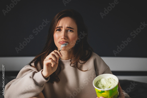 Breakup and sadness concept. Crying girl eating ice cream in bed at home, sad woman lick spoon with ice cream. Bad news, depressed woman. photo