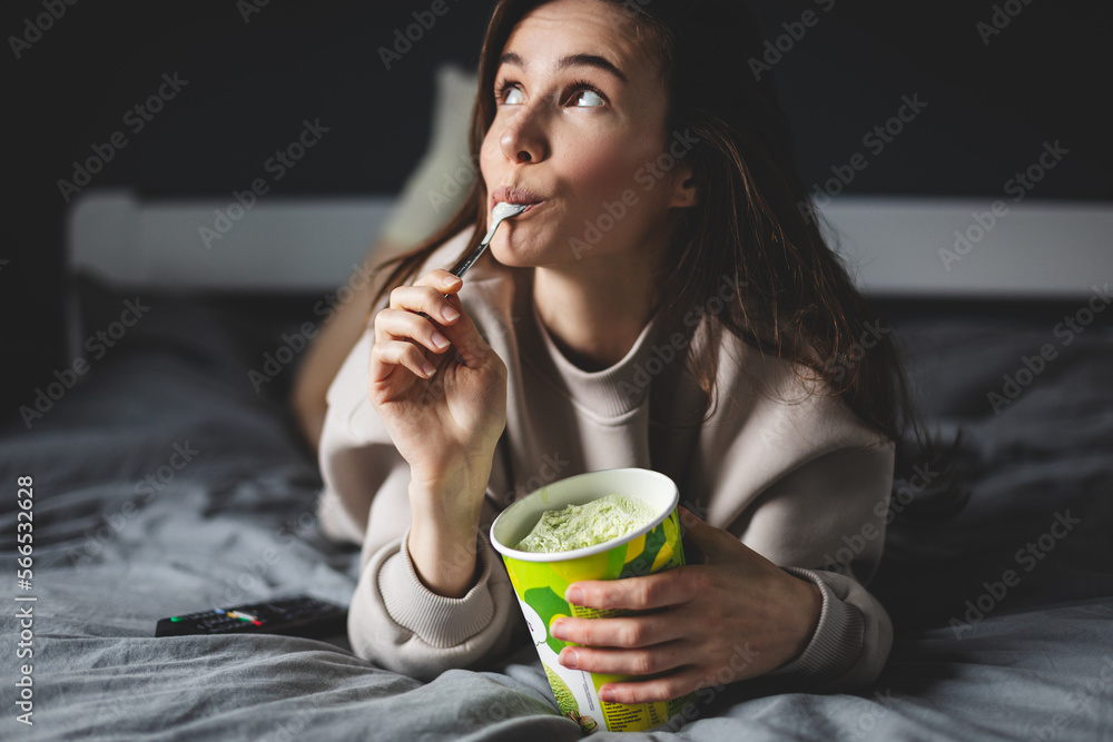 A young woman lies on her bed while eating a pint of pistachio ice cream with spoon. She is lick spoon with ice cream. Eating in bed. Happy beautiful woman resting in her comfortable bed at home.  - obrazy, fototapety, plakaty 