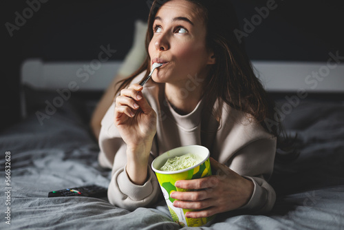 A young woman lies on her bed while eating a pint of pistachio ice cream with spoon. She is lick spoon with ice cream. Eating in bed. Happy beautiful woman resting in her comfortable bed at home.  photo