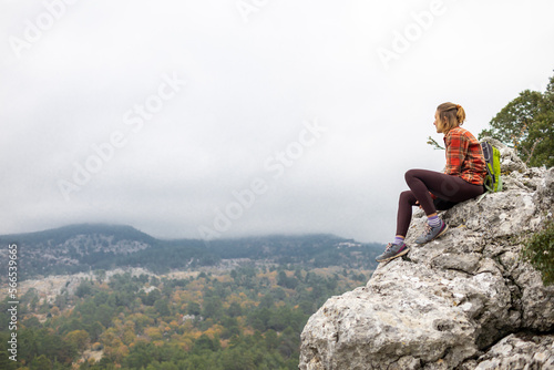 traveler girl sits on the edge of a cliff with an autumn forest and enjoys a beautiful view of the valley. © zhukovvvlad