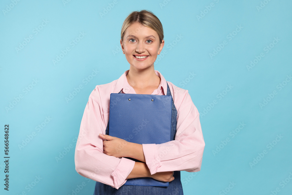 Beautiful young woman in denim apron with clipboard on light blue background Beautiful young woman in denim apron with clipboard on light blue background