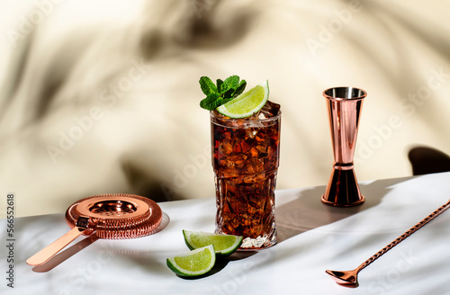 Whiskey cola cocktail with strong alcohol and ice, garnished with mint and lime in highball glass. Beige background, hard light