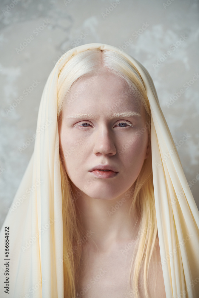 Fototapeta premium Young serene albino woman with white cover on head standing in front of camera against marble wall during photo session in studio