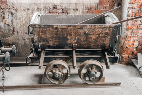 Old, rusted cart used in vintage factory or coal mine. Exhibition of industrial equipment. Metal trolley standing on rails. Rusted, brick wall in the background.