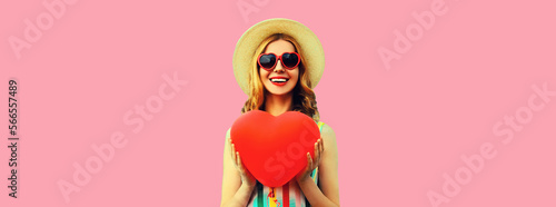 Portrait of happy smiling young woman with big red heart shaped balloon having fun wearing summer straw hat, sunglasses on pink background, blank copy space for advertising text © rohappy