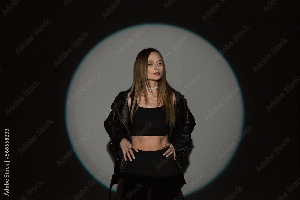 Fashionable young beautiful woman model in casual stylish black outfit with top and leather jacket stands on dark background with circle light in studio Fashionable young beautiful woman model in casual stylish black outfit with top and leather jacket stands on dark background with circle light in studio