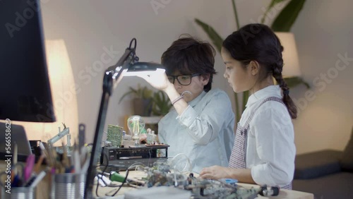 Two secondary school students doing project together. Cute girl and boy in glasses attaching details to electrical bulb set while boy pulling down desk lamp for better lighting. Education concept