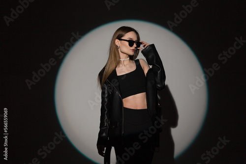 Stylish beautiful young hipster woman wears cool black sunglasses in fashion black rock clothes with top and leather jacket on dark background with hard circle light at the party