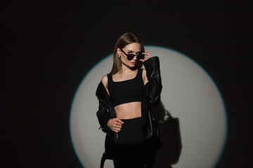 Fashionable beautiful stylish woman model hipster in trendy black rock clothes with top and fashion leather jacket puts on stylish sunglasses on a dark background with a round light