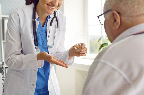 Friendly doctor gives a hearing aid to a senior patient with hearing loss. Happy smiling woman audiologist in scrubs and white medical coat gives an ear aid to a hearing impaired old man. Cropped shot photo