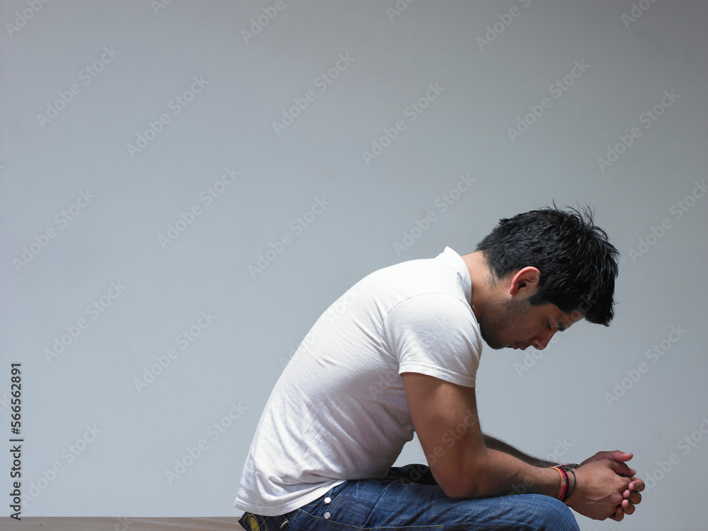 A Hispanic man dressed in a white T-Shirt and jeans sits with hands clasped and head down. A Hispanic man dressed in a white T-Shirt and jeans sits with hands clasped and head down.