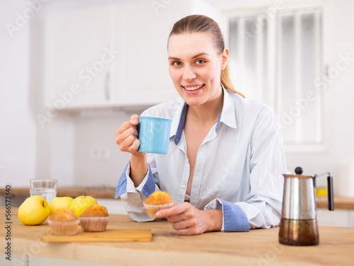 Foto Smiling young woman in male shirt drinking coffee in home kitchen