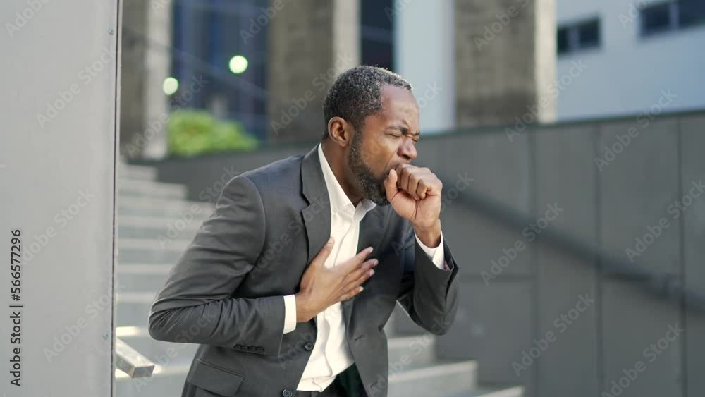 An african american mature man with a cold coughs while standing outside in front of an office building. Tired middle-aged businessman in formal suit holding his chest, suffering from virus, flu An african american mature man with a cold coughs while standing outside in front of an office building. Tired middle-aged businessman in formal suit holding his chest, suffering from virus, flu