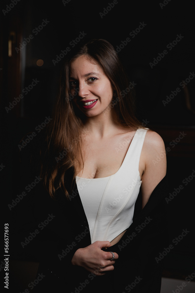 Fototapeta premium The portrait of young sensual brunette caucasian woman in white top, black jacket and bright make up smiling, dark background