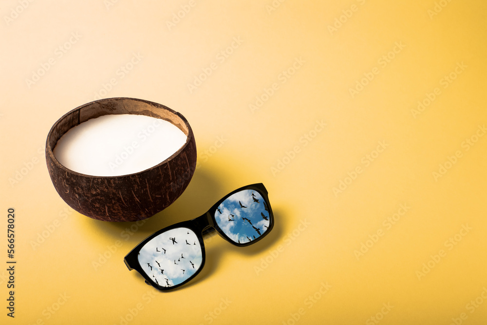 Summer composition on the theme of vacation. Coconut with sunglasses on a yellow background. Reflection of birds flying in the blue sky in glasses lenses. Copy space Summer composition on the theme of vacation. Coconut with sunglasses on a yellow background. Reflection of birds flying in the blue sky in glasses lenses. Copy space