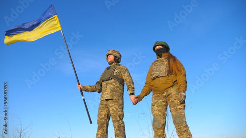 Male and female ukrainian army soldiers holding hands of each other and lifted up flag against blue sky. Military people waving flag of Ukraine in honor of the victory against Russian aggression