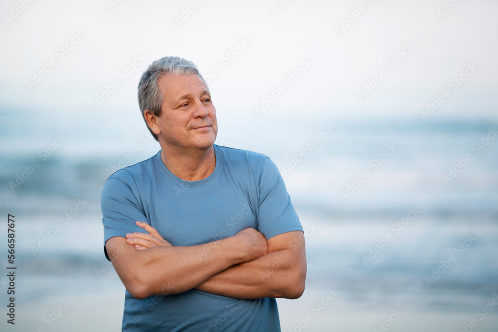 Middle shot of a smiling senior man looking away with arms crossed on the chest. Cheerful person on blurry background of sea and sky