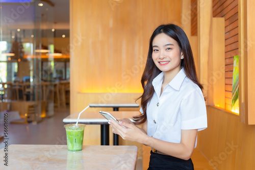 Portrait of cute an Asian Thai girl student in uniform is sitting smiling happily while holding smartphone in a coffee shop in university.