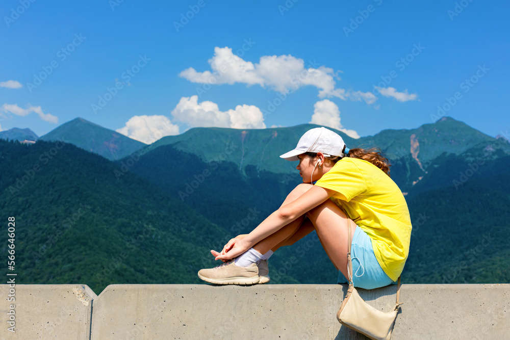 A teenage girl in a yellow T-shirt and baseball cap sits on the parapet overlooking a mountainous landscape in the summer. The concept of solitude, teenage detachment.  