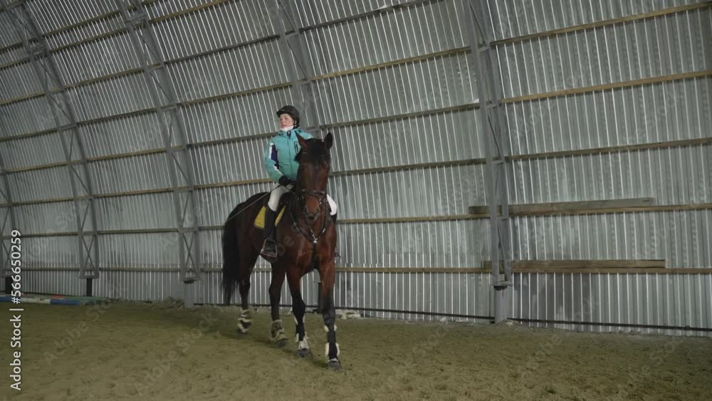 A woman rides a horse indoors. Young woman doing horseback riding in indoor manege. A woman rider on a horse in a winter hangar. A woman rides a horse indoors. Young woman doing horseback riding in indoor manege. A woman rider on a horse in a winter hangar.