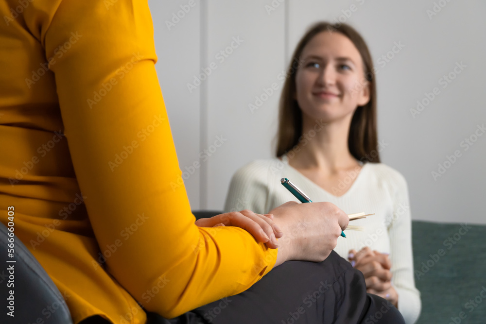 Focus on the psychologist's hand with a pen. Smiling young woman listens to a psychologist at the reception, sitting on the couch in the office.Therapist consults the patient.