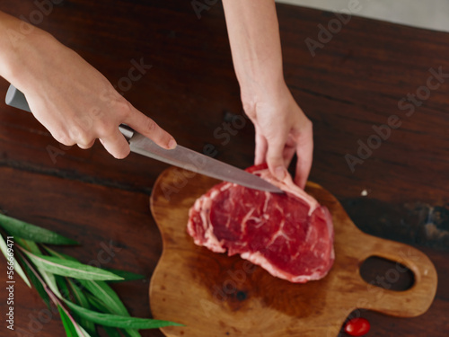 Woman with knife in hand cutting fresh steak meat for roasting kitchen with salt pepper other spices on table, red cherry tomatoes and herbs, preparing dinner in home kitchen. wooden table, top view.