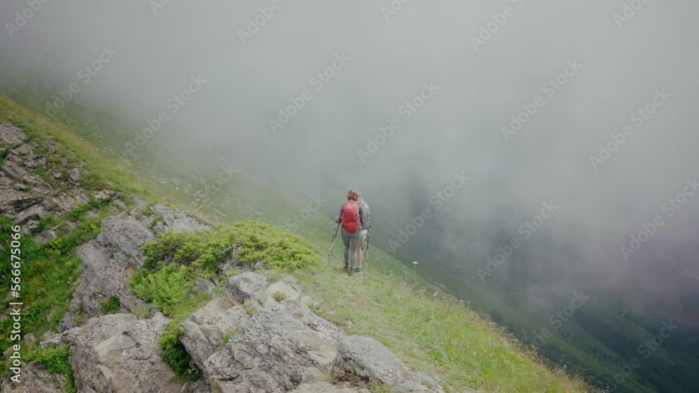 Two women with backpacks and trekking poles hiking in mountains amidst meadows Two women with backpacks and trekking poles hiking in mountains amidst meadows
