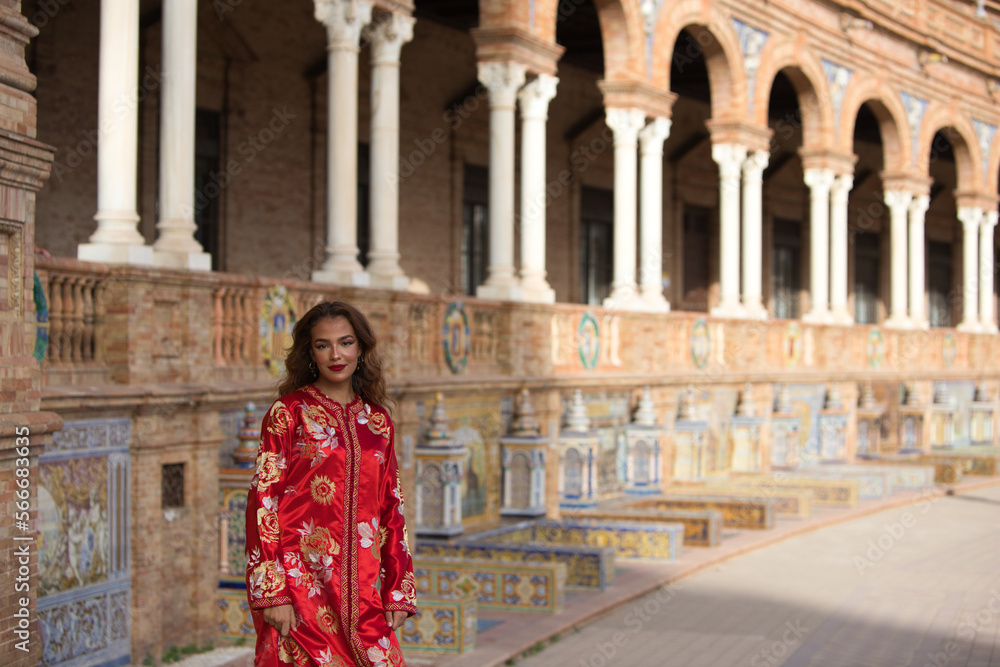 A beautiful young woman wearing a traditional Moroccan red dress with gold and silver embroidery is on vacation in Seville, Spain.