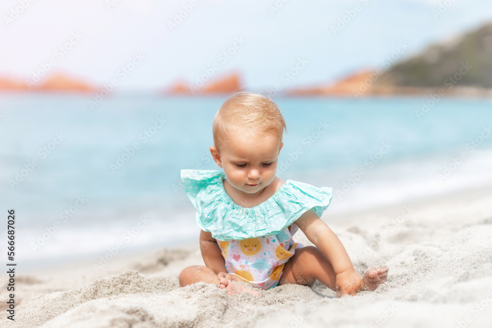 Beautiful baby girl playing with sand while sitting in a swimsuit on the beach in summer. Summer weekend, family travel, vacation concept Beautiful baby girl playing with sand while sitting in a swimsuit on the beach in summer. Summer weekend, family travel, vacation concept