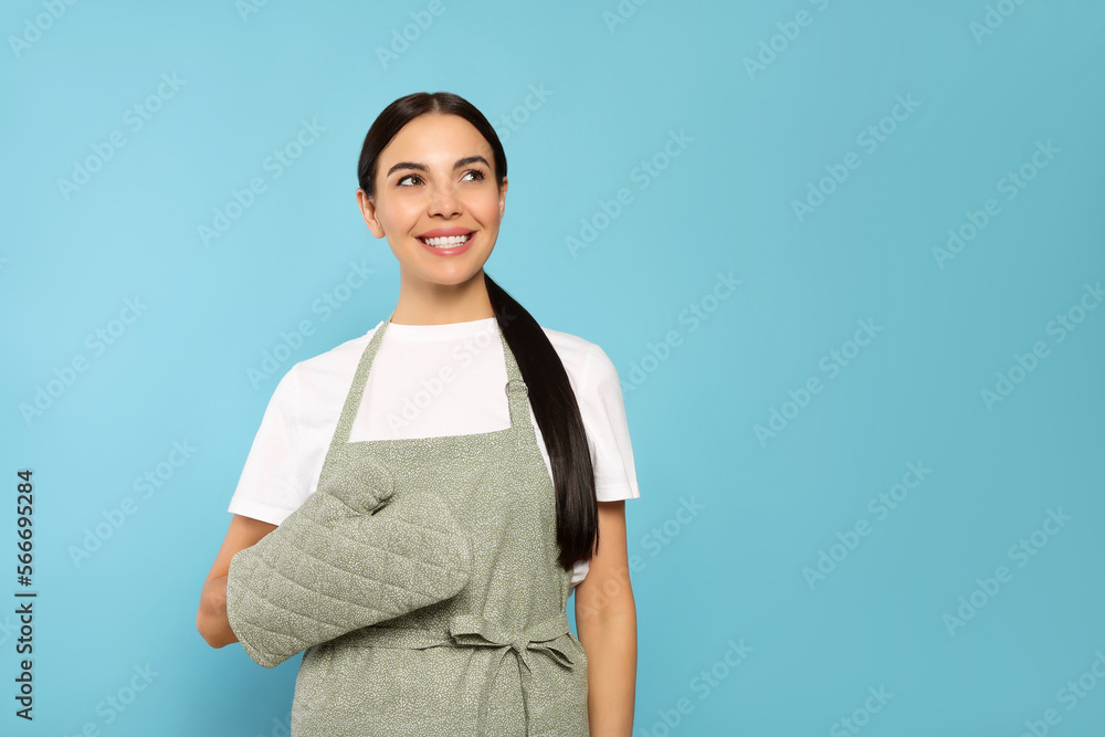 Young woman in grey apron and oven glove on light blue background, space for text Young woman in grey apron and oven glove on light blue background, space for text