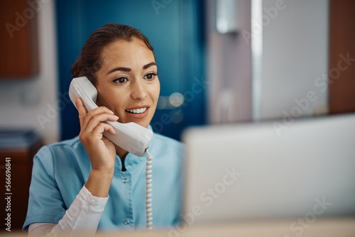 Young nurse answering phone call while working on desktop PC at reception desk in hospital.