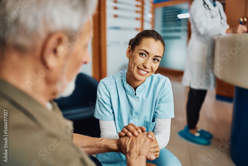 Caring nurse holding hands with senior patient while talking to him in waiting room at doctor's office.