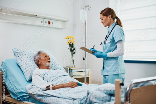 Nurse practitioner writes data in medical record of hospitalized senior patient while visiting him during recovery at intensive care unit.