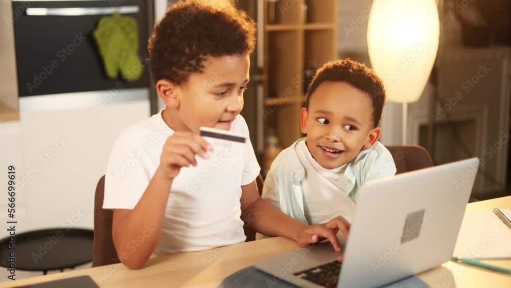 Two cute little african american boys brothers paying with credit card on laptop computer at home Happy young kids customers doing payments online shopping buy toys in internet store Two cute little african american boys brothers paying with credit card on laptop computer at home Happy young kids customers doing payments online shopping buy toys in internet store