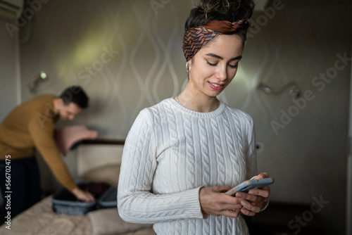 couple young beautiful woman and man in hotel room wife or girlfriend use mobile phone for texting sens sms messages while husband or boyfriend is unpacking or packing baggage on the bed copy space
